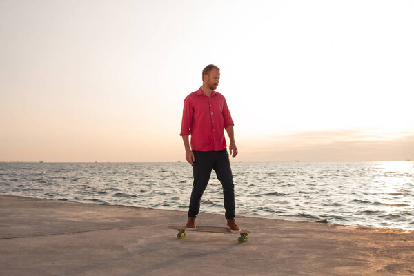 skater in red shirt and blue jeans riding near beach on longboard during sunrise, sea or ocean background
