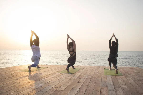 Group of people in yoga poses stand on the wooden pier, sunrise in the sea background