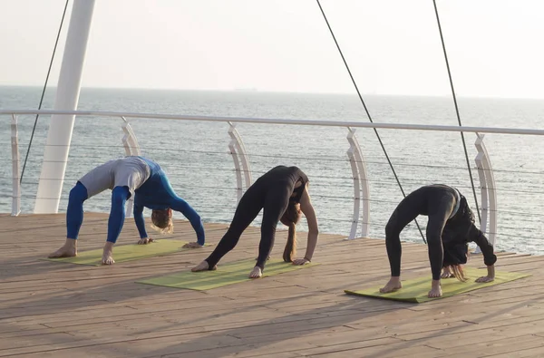 Group of people in yoga poses stand on the wooden pier, sunrise in the sea background