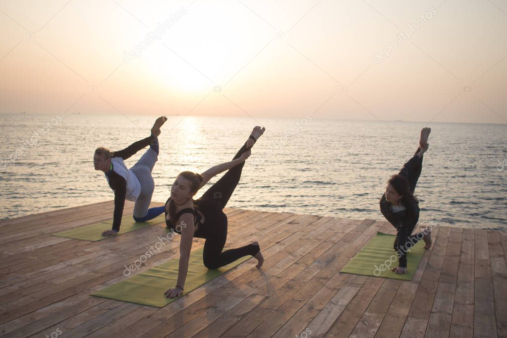 Group of people in yoga poses stand on the wooden pier, sunrise in the sea background