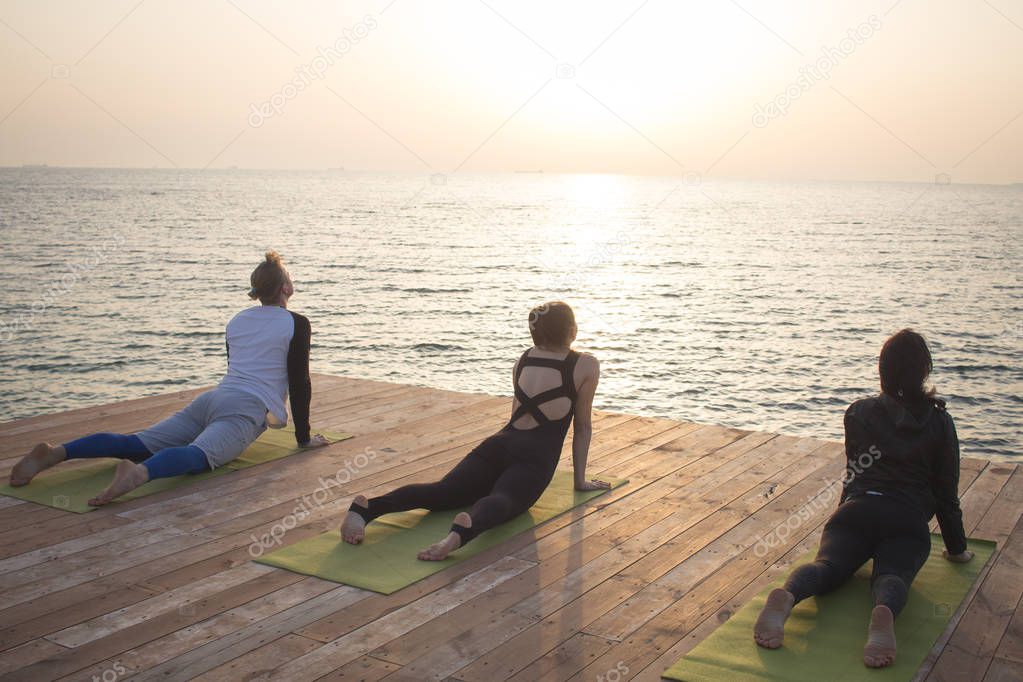 Group of people in yoga poses stand on the wooden pier, sunrise in the sea background