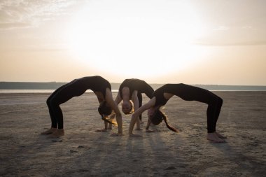 Genç kadınlar Yoga uygulamak pozlar ve asanas. Ortak yoga akrobatik yoga. Çölde siyah giymek eğitim gün batımı sırasında yoga sınıfında