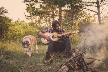 Halk veya ülke gitar çalan köpek sahip, adam woods, kamp ateşi ve gün batımı arka plan