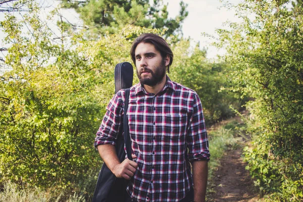 Young bearded male with long hair and guitar, portrait of traveler musician hiking in the forest