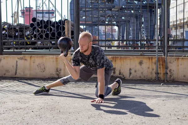 Young bearded male athlete training in industrial zone in sunny day, kettlebells exercises outdoors, urban background