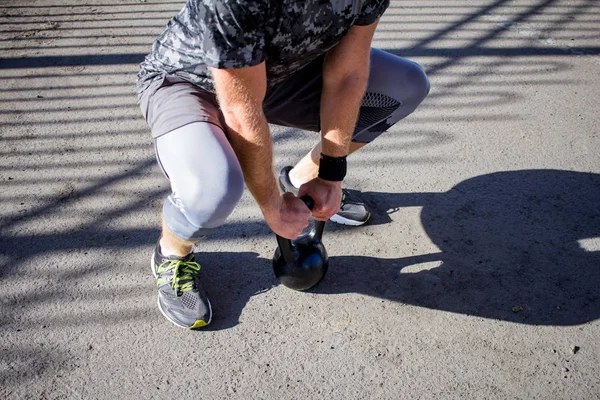 Young bearded male athlete training in industrial zone in sunny day, kettlebells exercises outdoors, urban background