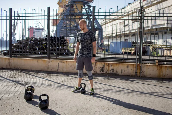 Young bearded male athlete training in industrial zone in sunny day, kettlebells exercises outdoors, urban background