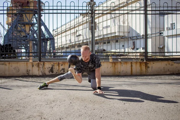 Young bearded male athlete training in industrial zone in sunny day, kettlebells exercises outdoors, urban background