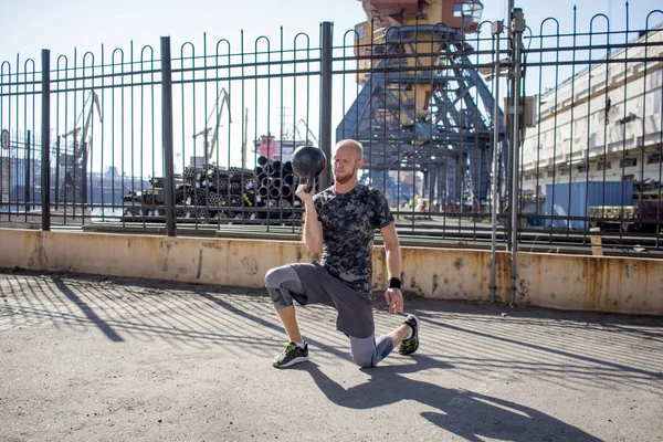 Young bearded male athlete training in industrial zone in sunny day, kettlebells exercises outdoors, urban background