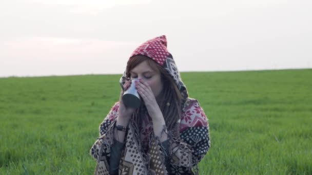 コーヒー ポットとポンチョで若い女性の緑のフィールドでドレッドヘアを持つ赤毛の女性が熱いお茶を飲む — ストック動画