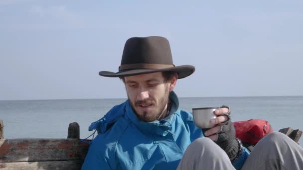 ブルーのジャケットと帽子ドリンク コーヒーまたは紅茶ビーチ 古い木造船の背景上でハイカーにひげを生やした若い — ストック動画