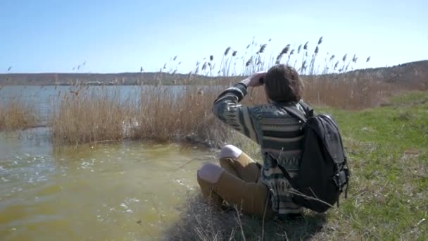 Young male with backpack and binoculars relaxing near the river in sunnyday — Stock Footage Young Male Backpack Binoculars Relaxing River Sunnyday Stock Video