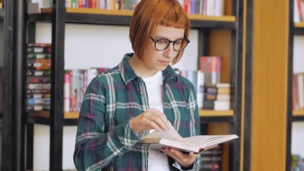 Молодая Рыжая Женщина Очках Читает Книги Библиотеке — стоковое видео