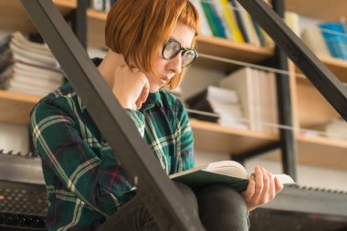 Gözlük Genç Redhead kadın kütüphanede kitap okumak