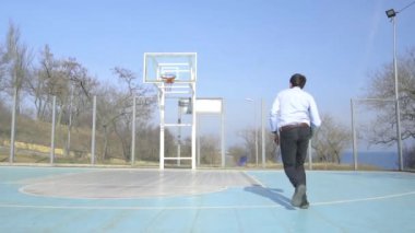 Takım elbiseli ve kravatlı genç iş adamı güneşli bir günde açık havada tek başına basketbol oynuyor