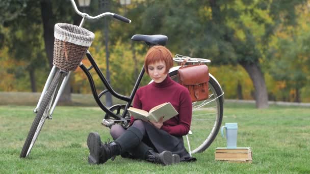 秋天公园里的年轻女子读书 美丽的红头发的女伴着自行车在绿草上 — 图库视频影像