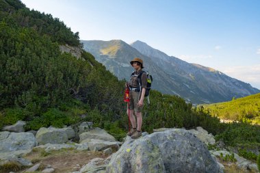 Sırt çantası ve gaz feneriyle güzel dağlarda duran genç ve mutlu bir kadın.