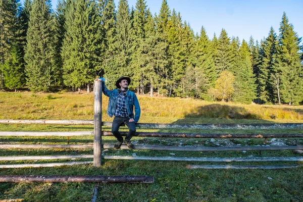 身穿粗斜纹棉布夹克头戴牛仔帽的年轻男性农民站在木栅栏旁边 挡住森林 — 图库照片