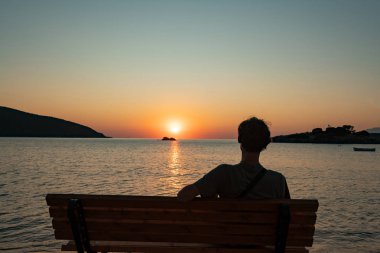 Erkek silueti bankta oturuyor ve yaz kumsalında güneşin doğuşunu seyrediyor.