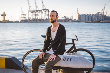 Günbatımı sırasında deniz bağlantı noktası arka plan üzerinde oturan, bir kenara görünümlü şık hipster adam