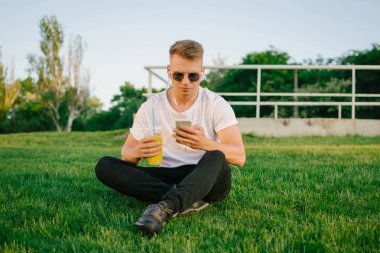 Yakışıklı genç hipster adam akıllı telefon açık havada park limonata el ile kullanarak