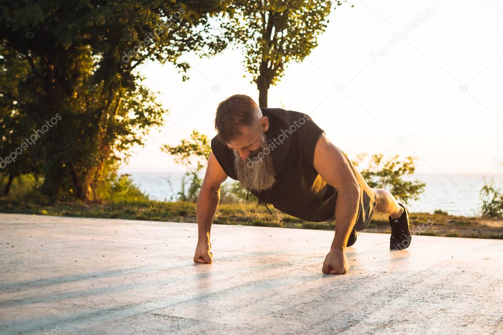 Handsome middle aged man with long gray beard doing pushups during sunrise