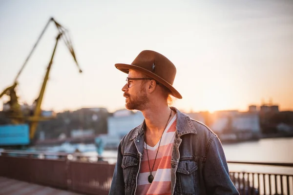 Young stylish hipster in round glasses and hat on sea port background during sunset