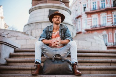 Genç şık yakışıklı redbeard adam çanta ile ve eski şehir merkezinde merdivenlerde oturan şapkalı