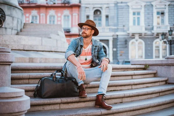 Genç şık yakışıklı redbeard adam çanta ile ve eski şehir merkezinde merdivenlerde oturan şapkalı
