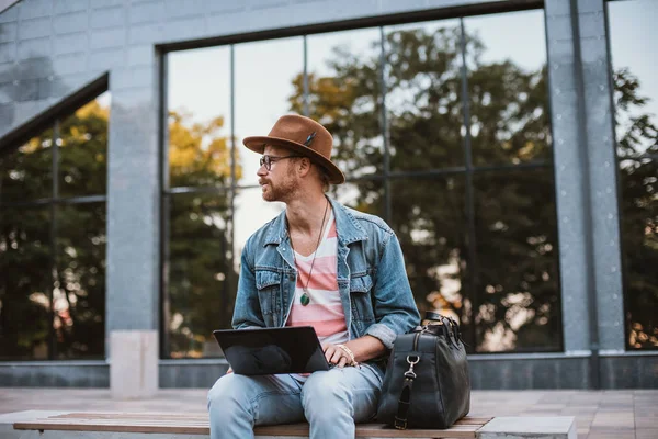Young bearded hipster man with bag and in hat sits on bench in city center in park using laptop
