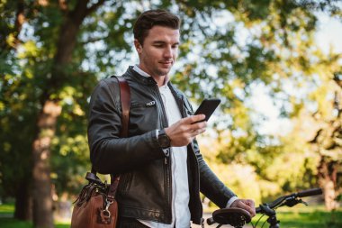 Yakışıklı genç şık adam akıllı telefon açık havada Bisiklet Park ile kullanma. 