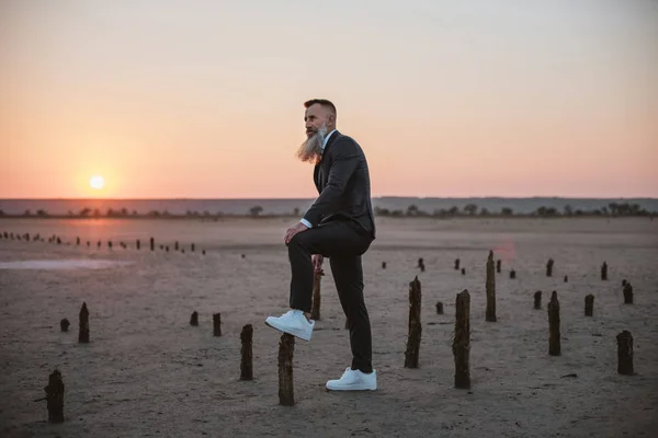 Handsome bearded mature man in suit posing outdoors on beach during sunset