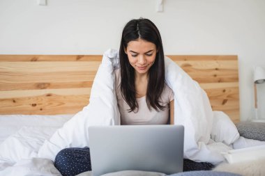 Evde battaniye ile kaplı yatakta dizüstü bilgisayar kullanan neşeli genç kadın
