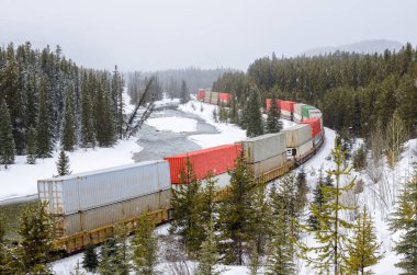 Cargo Train Running along a Frozen Mountain River during heavy Snowfall on a Winter Day. Banff National Park, AB, Canada. clipart