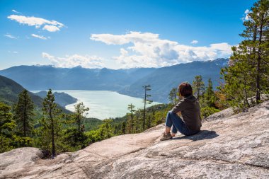 Kadın uzun yürüyüşe çıkan kimse fiyort çevrili Wooded dağlar tarafından hayran bir dağın tepesinde bir kayanın üzerine oturmuş. Squamish, Bc, Kanada.