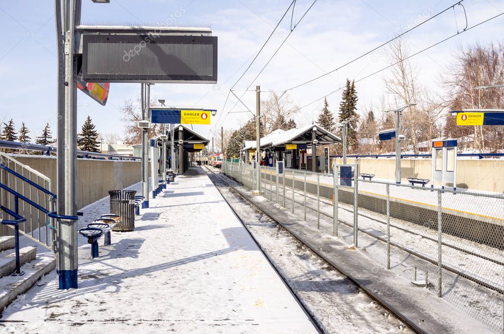 Deserted Platform at a Light Rail Station Covered in Snow on a Sunny Winter Day. An Approaching Train is Visible in Background. Calgary, AB, Canada.