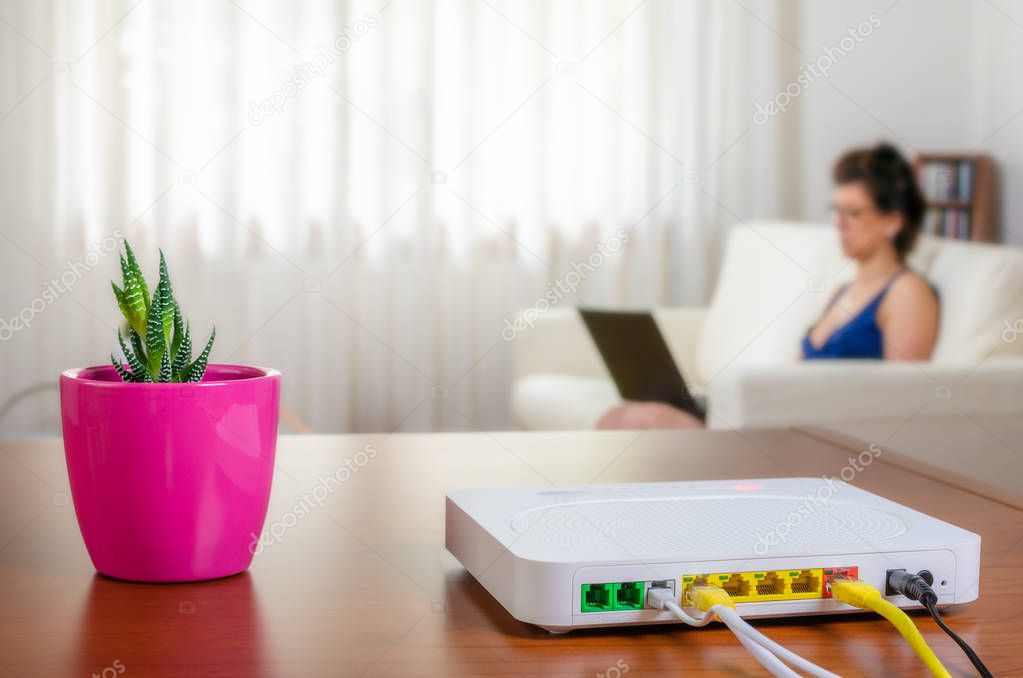 Close view of a modem router on a wooden table in a living room with a woman using a laptop while sitting on a sofa in background