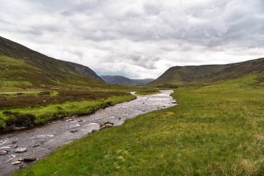 Bulutlu bir yaz gününde dolambaçlı bir dağ nehri. Nehir boyunca bir yol akar. Dee Nehri, Aberdeenshire, İskoçya, Uk.