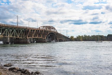 Yarı bulutlu bir sonbahar gününde eski paslı bir çelik yol köprüsünün altından geçen büyük bir nehirde yelken açan tekneler. Portsmouth, NH, ABD.