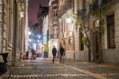 İnsanlar geceleyin eski bir şehir merkezinde tarihi binalarla dolu kaldırım taşı kaldırımlı bir caddede geziniyorlar. Antwerp, Belçika.