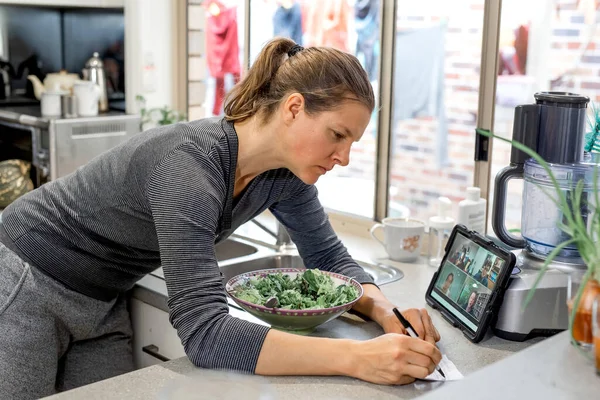 Mujer en la cocina viendo zoom-reunión en la tableta y escribe notas — Foto de Stock