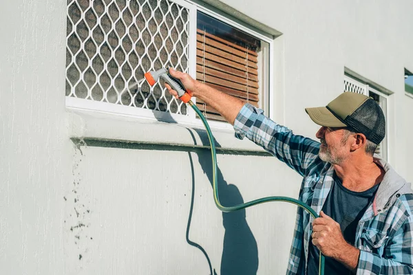 El hombre limpia la ventana con el flujo de agua por tubo de manguera Imágenes de stock libres de derechos