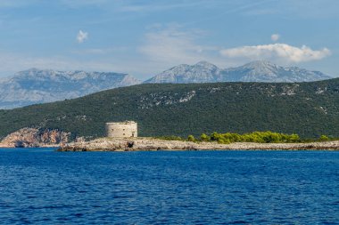 Taşlı deniz kıyısı, ufukta dağlar ve yuvarlak kale, Karadağ