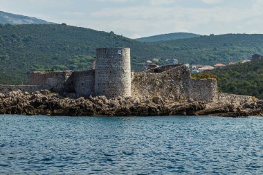 Taşlı deniz kıyısı, ufukta dağlar ve eski yuvarlak kale