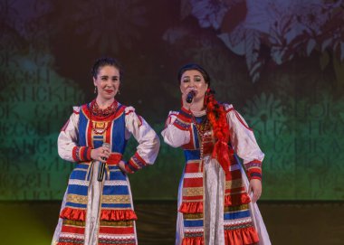 Parlak ulusal kostümlü kızlar sahnede şarkı söyleyip dans ediyorlar.