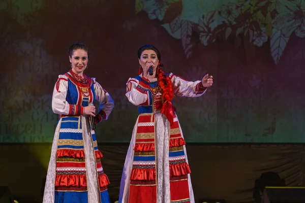 Parlak ulusal kostümlü kızlar sahnede şarkı söyleyip dans ediyorlar.