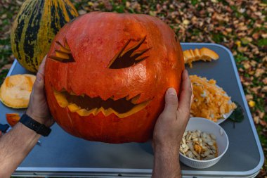 Hands hold a carved pumpkin with a spooky face in a fall setting. Nearby, other pumpkins and pumpkin guts are scattered on a table covered in autumn leaves.