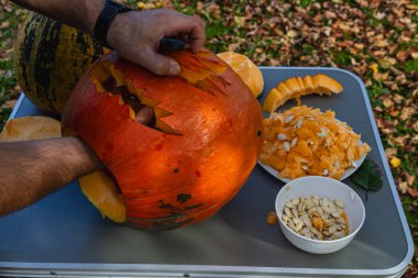Hands skillfully carve a spooky face into a bright orange pumpkin surrounded by fresh pumpkin guts and seeds on a table outdoors in autumn leaves.