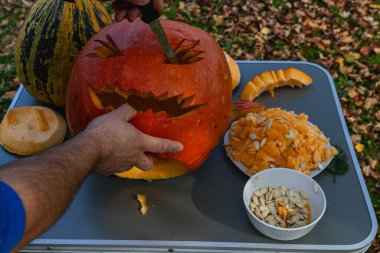 Hands carefully carve a scary face into a bright orange pumpkin surrounded by autumn leaves. Nearby, pumpkin seeds and flesh are neatly arranged on a table.