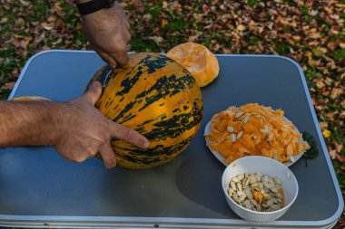 Hands are seen skillfully cutting into a striped pumpkin on a table amidst fallen leaves, with pumpkin flesh and seeds arranged in separate bowls nearby.